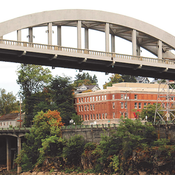 A large arched bridge spans over a lush, tree-filled area with a red brick building in the background.